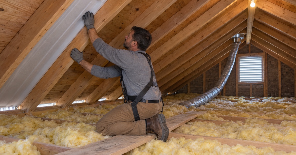 A roofer in the attic of a 1970s Waterford house, fitting a vent baffle at the eave so the soffit vents can breathe.
